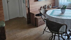 Uma sala de jantar com uma mesa redonda, cadeiras de vime, uma TV de tela plana na parede e uma janela com cortinas.