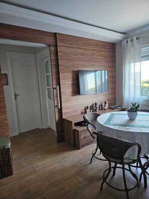 A dining area with a round table, wicker chairs, a flat-screen TV mounted on the wall, and a window with curtains. - Apartment on the beachfront next to the Aquarium. (Balneário Camboriú)