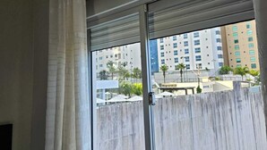 Quarto com uma grande janela, cortina e vista para um horizonte urbano com edifícios e palmeiras.