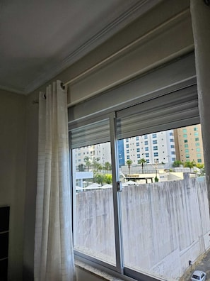 A room with a large window, a curtain, and a view of a cityscape with buildings and palm trees. - Apartment on the beachfront next to the Aquarium. (Balneário Camboriú)