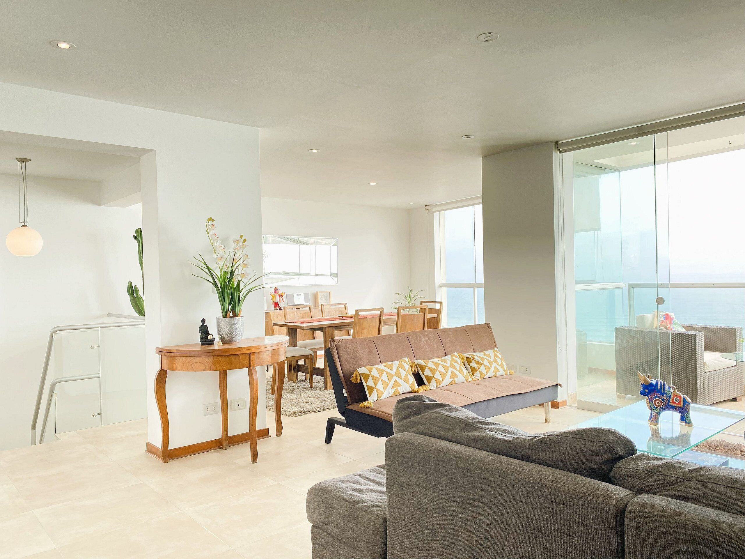 A modern living room with a sofa, a dining table, and chairs. There is a balcony with a glass railing.