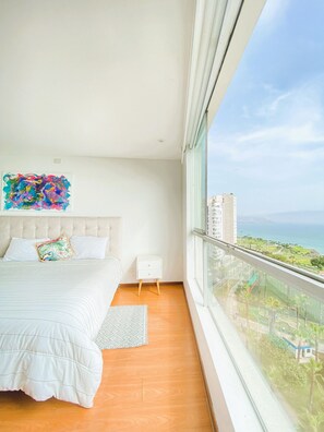 A bedroom with a large bed, a nightstand, a colorful abstract painting, and a view of a cityscape and ocean. - Miraflores duplex with ocean view garage (Lima)