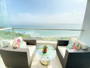 A balcony with wicker furniture and a glass table, overlooking a coastal view. - Miraflores duplex with ocean view garage (Lima)