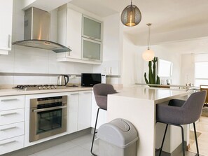A modern kitchen with stainless steel appliances, a built-in oven, a microwave, and a coffee maker. There is a large island with bar stools and a trash bin. - Miraflores duplex with ocean view garage (Lima)