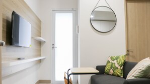 A living room with a flat-screen TV mounted on the wall, a round mirror, a sofa with patterned cushions, and a small table. - Pleasurable And Modern 2Br Tokyo Riverside Pik 2 Apartment (Teluknaga)