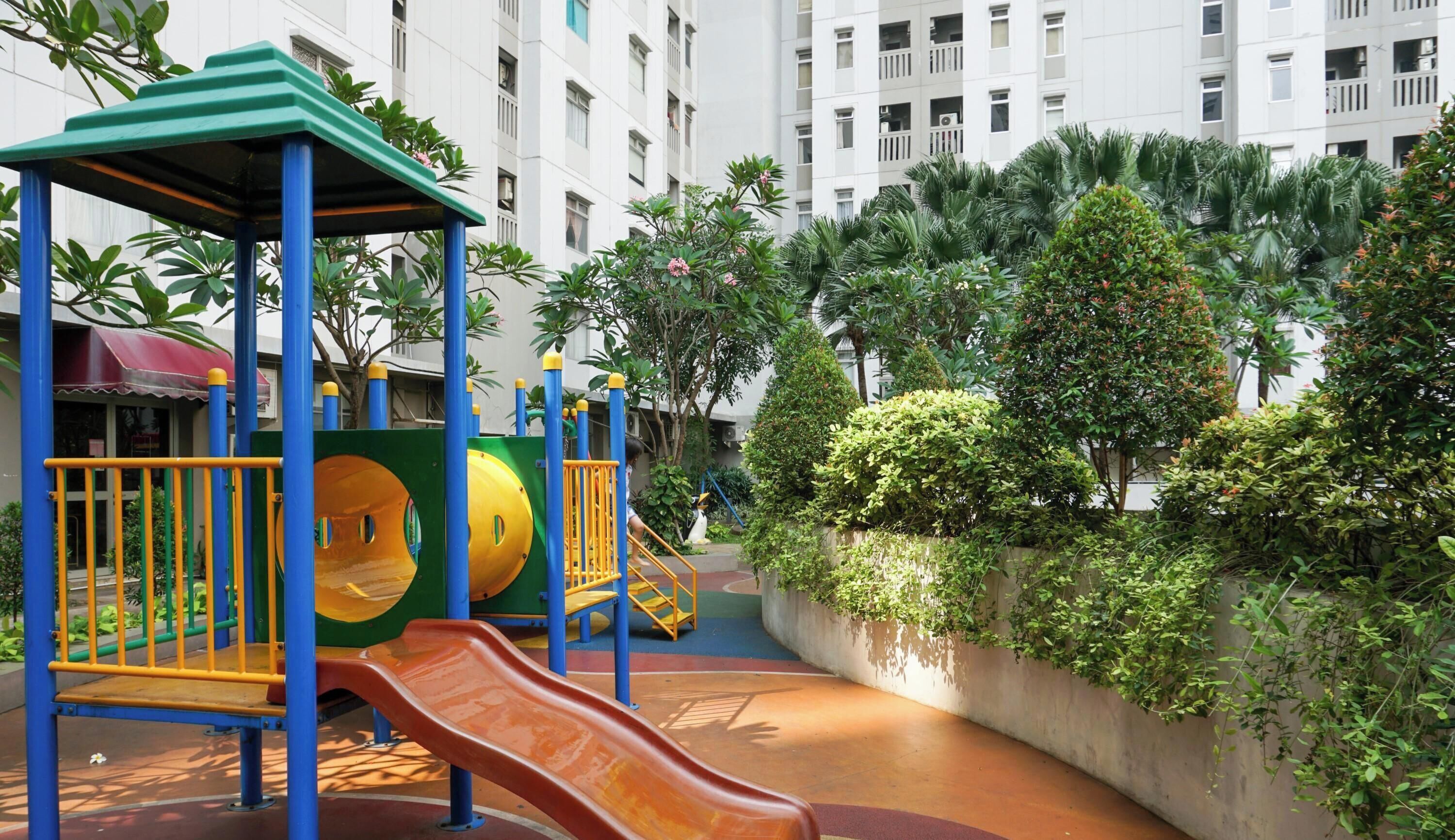 Un parque infantil con un tobogán y estructuras de escalada, rodeado de vegetación y un edificio al fondo.