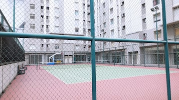 Una cancha de tenis al aire libre rodeada por una cerca de malla metálica, con un edificio de varios pisos al fondo.