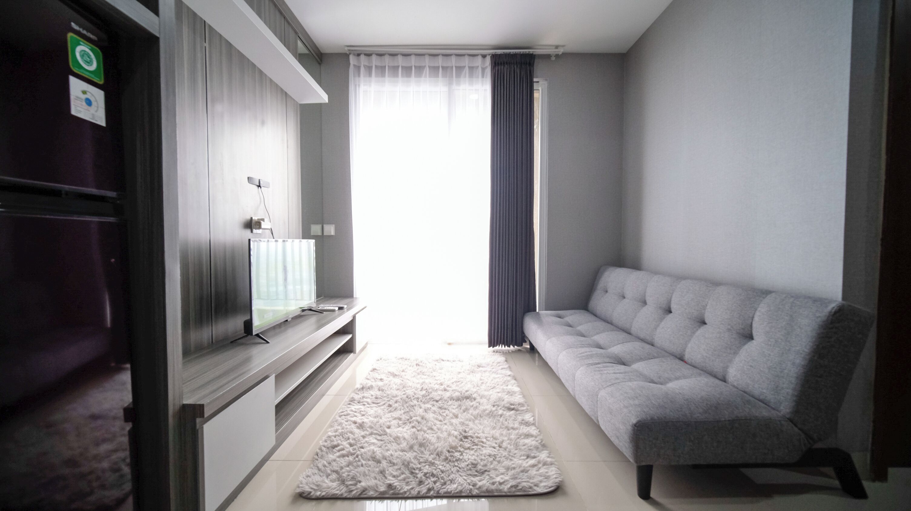 Ein modernes Wohnzimmer mit einem grauen Sofa, einem Flachbildfernseher, einem flauschigen Teppich und einem Fenster mit Vorhängen.