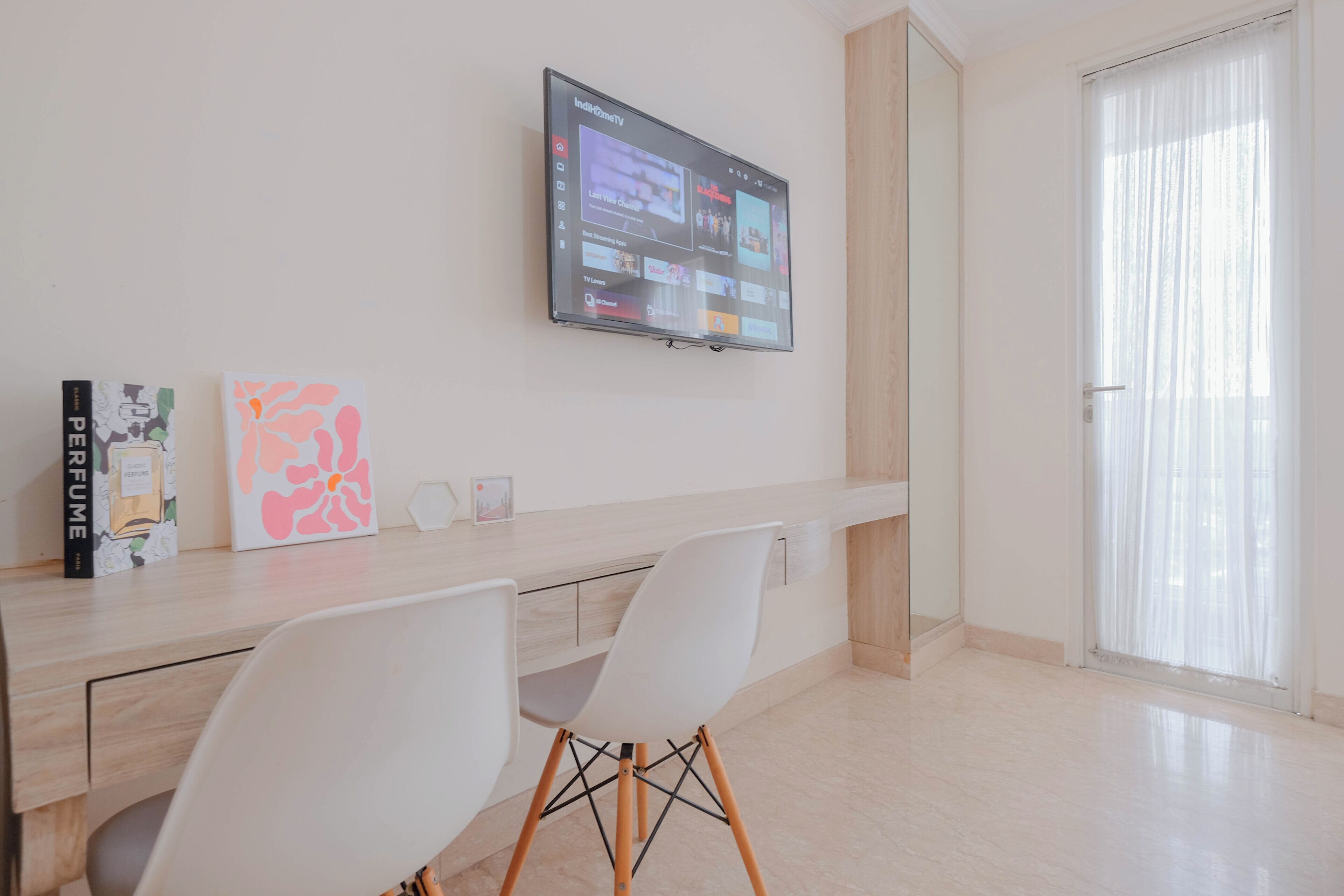 A modern living room with a flat-screen TV mounted on the wall, a wooden dining table with two white chairs, and a large window with sheer curtains.