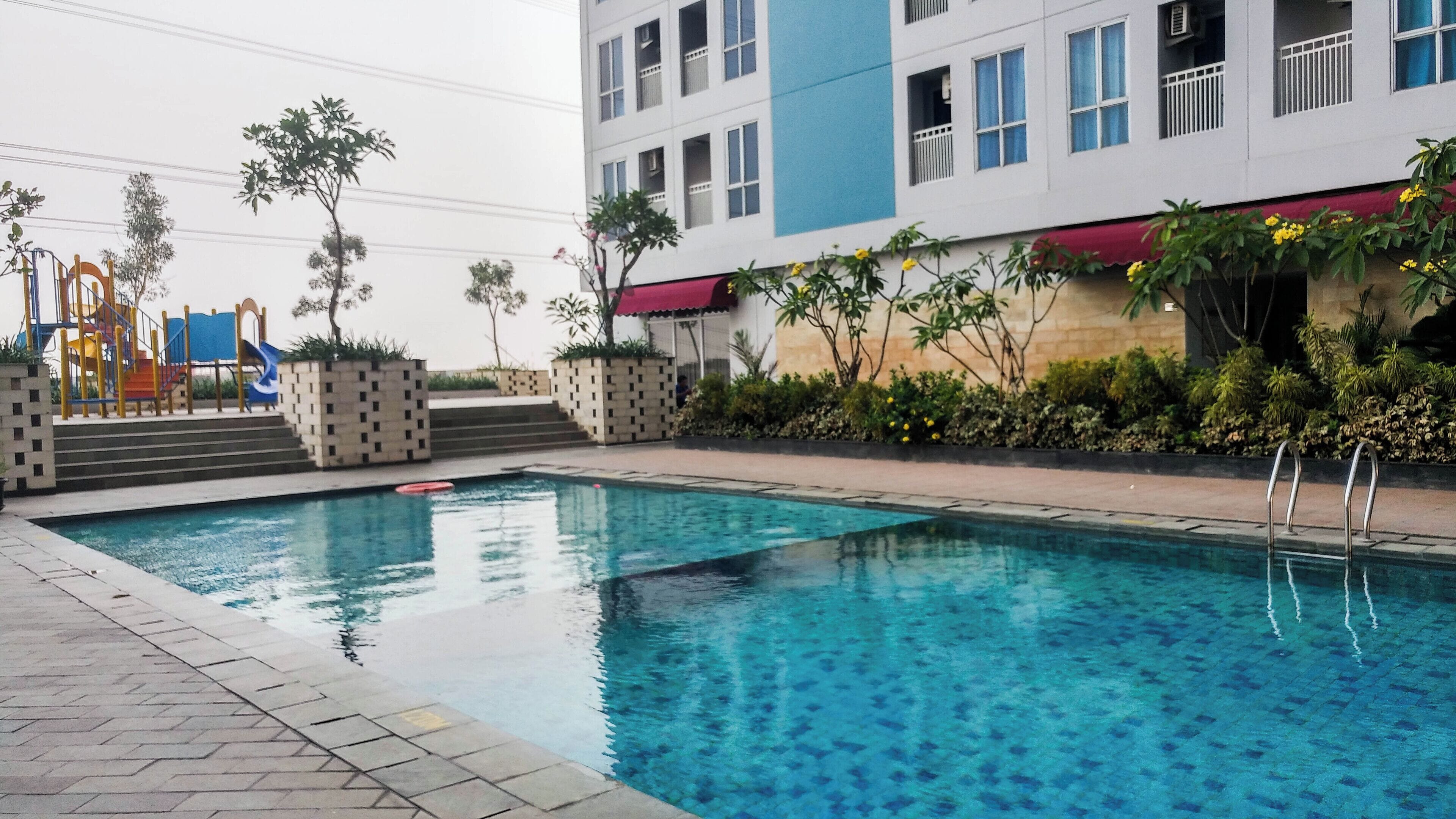 Una piscina con superficie de agua azul cristalina, rodeada por un sendero empedrado. Se observan escaleras que conducen a un edificio de fachada azul y blanca, y un parque infantil con juegos de colores al fondo.