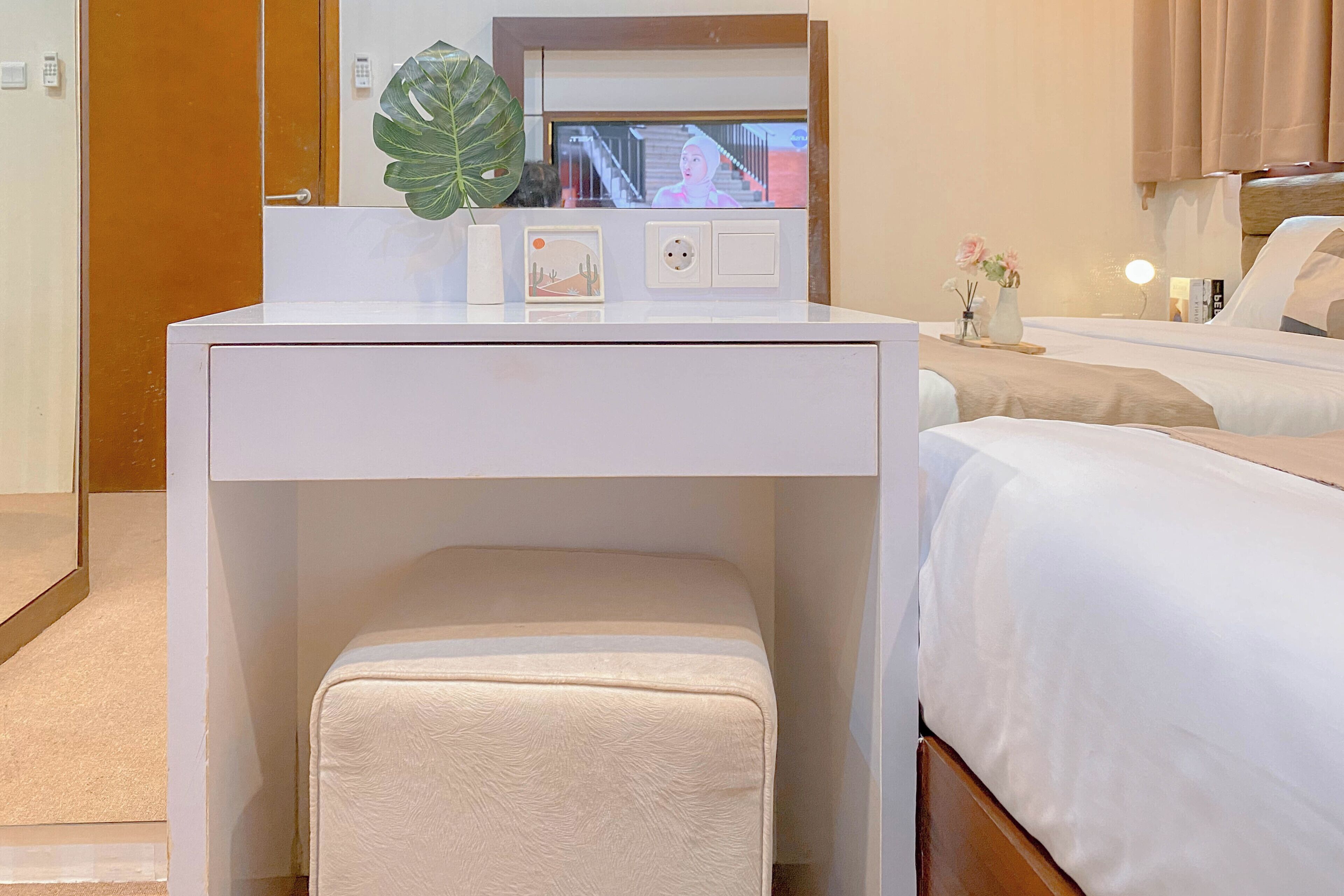 Une chambre d’hôtel avec un bureau blanc, un petit banc, un lit avec une literie blanche, un miroir et une plante posée sur le bureau.