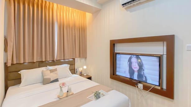 A hotel room with a bed, a TV mounted on the wall, a bedside table with a lamp, and curtains. - Minimalist And Good Place 1Br At Signature Park Grande Apartment (Jakarta)