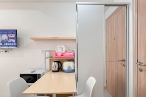 A room with a wooden table, a kettle, a microwave, a pink tray, and a television displaying a program. - Restful Living Studio (No Kitchen) At Apartment Bandaraya - Tallasa City Makassar (Makassar)
