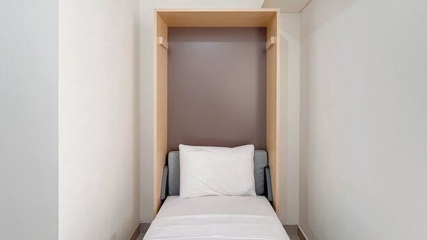 A narrow room with a bed, a wooden wardrobe, and a grey chair. - Restful Living Studio (No Kitchen) At Apartment Bandaraya - Tallasa City Makassar (Makassar)