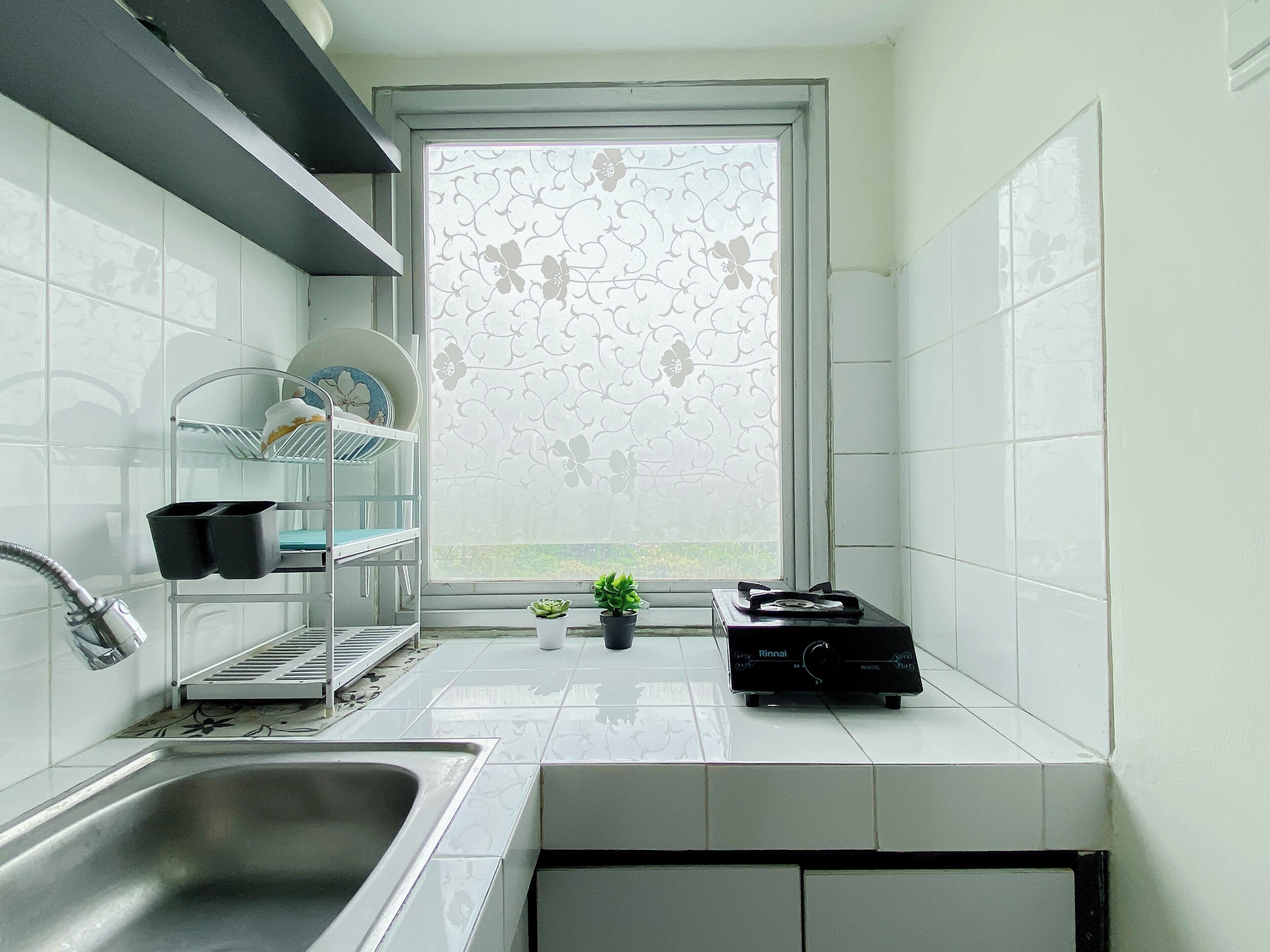 A modern kitchen with a stainless steel sink, a window with a floral pattern, and a shelf with dishes.