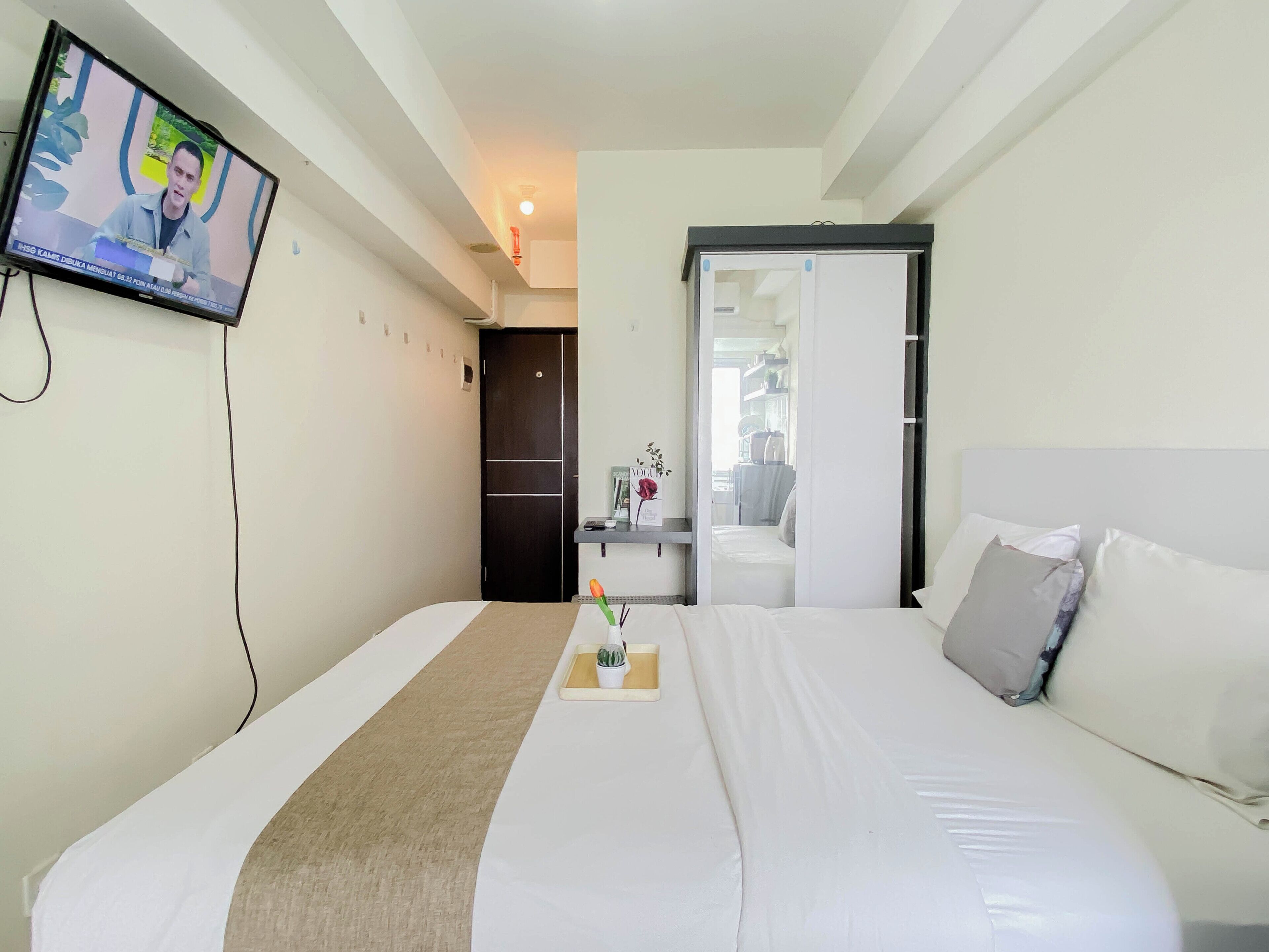 Una camera d'hotel con un letto, un comodino con un vaso e una pianta, una televisione a parete e una porta che dà su un'altra stanza.