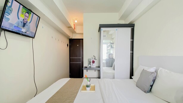 Habitación de hotel con cama, mesita de noche con un florero y una planta, televisor montado en la pared y puerta que da a otra habitación.