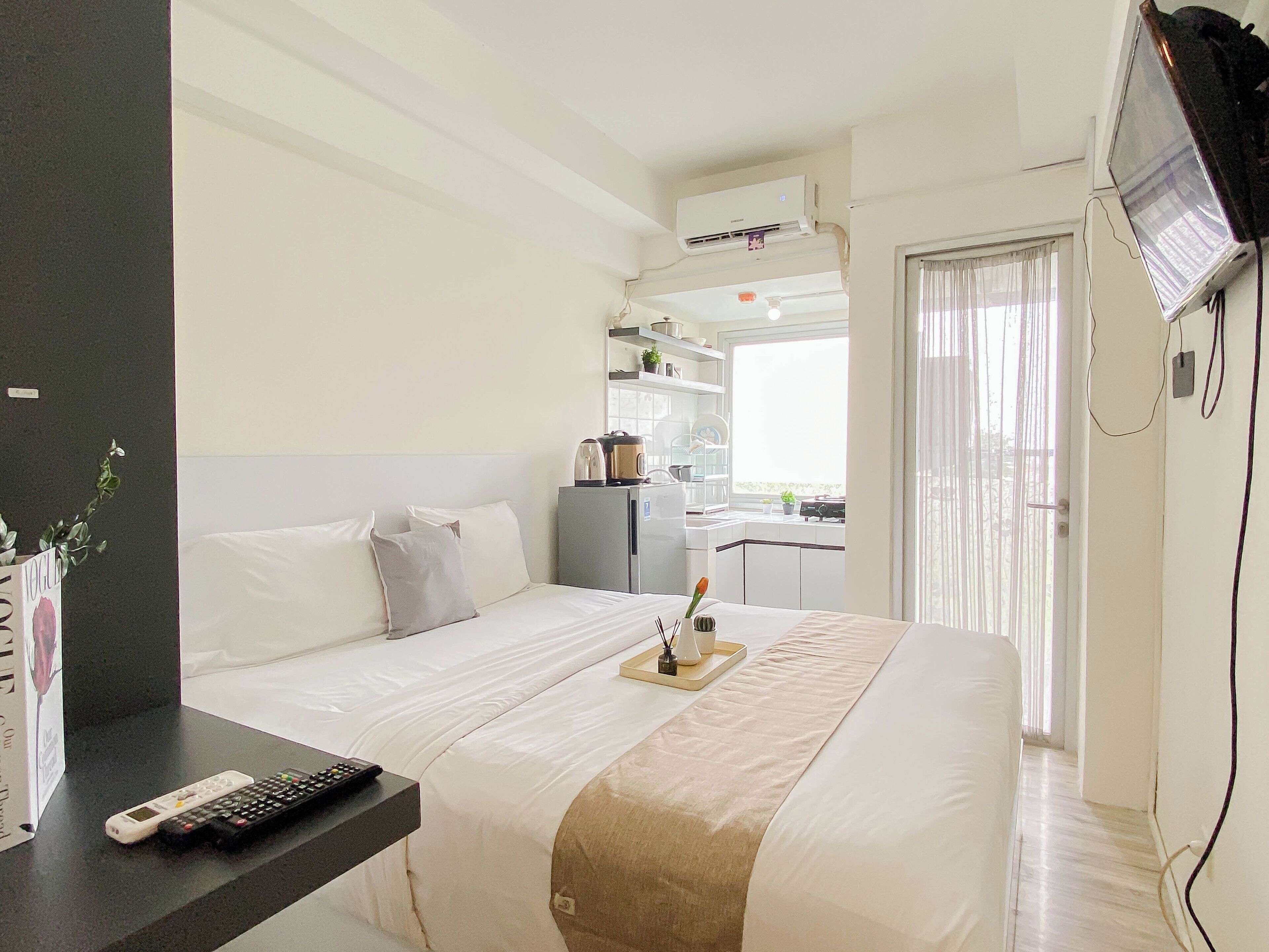 A hotel room with a bed, a desk with a remote control, a TV mounted on the wall, and a kitchenette area.