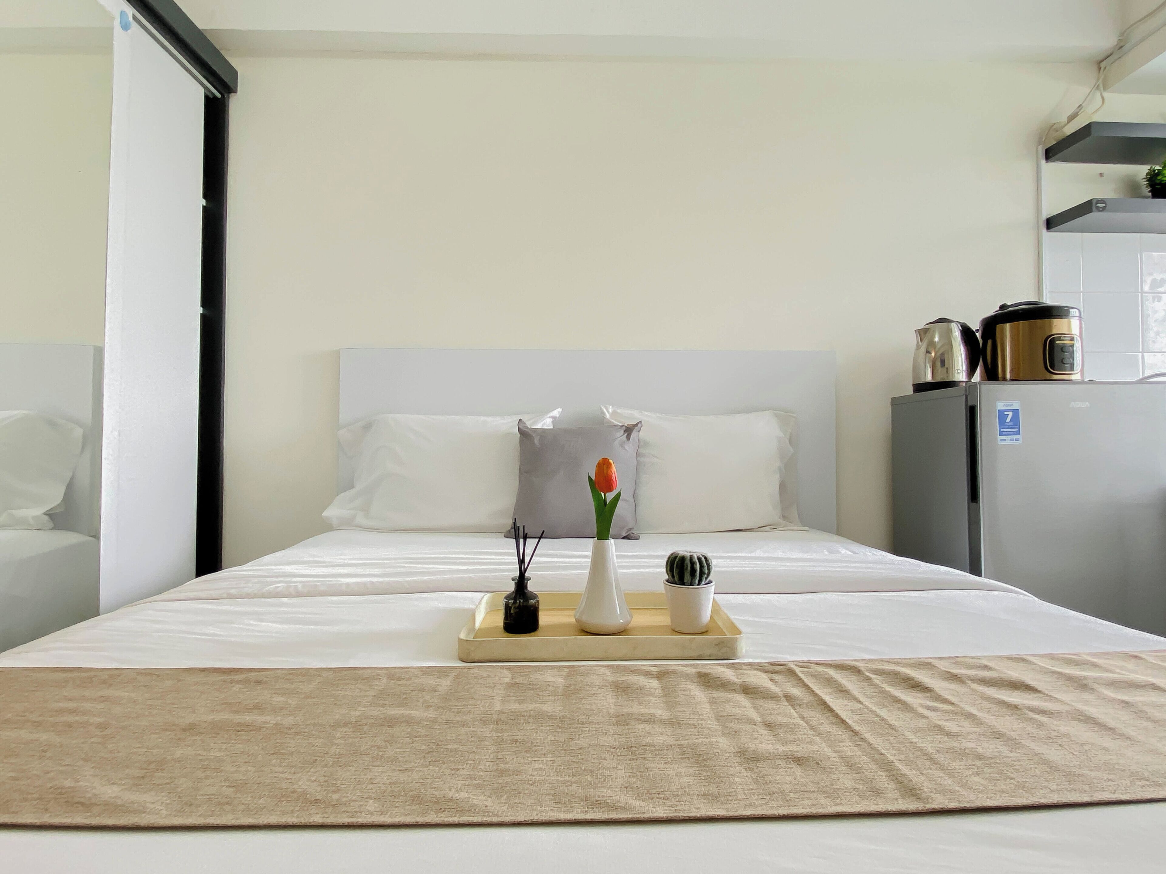 Quarto de hotel com cama, mesa de cabeceira com um vaso e uma vela, uma pequena planta e um frigorífico.