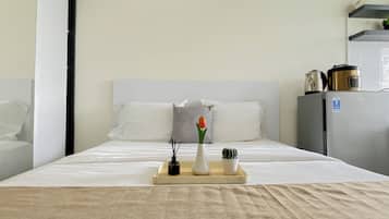 A hotel room with a bed, a bedside table with a vase and a candle, a small plant, and a refrigerator.