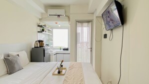 Habitación de hotel con cama, televisor montado en la pared, zona de kitchenette y ventana con cortinas.