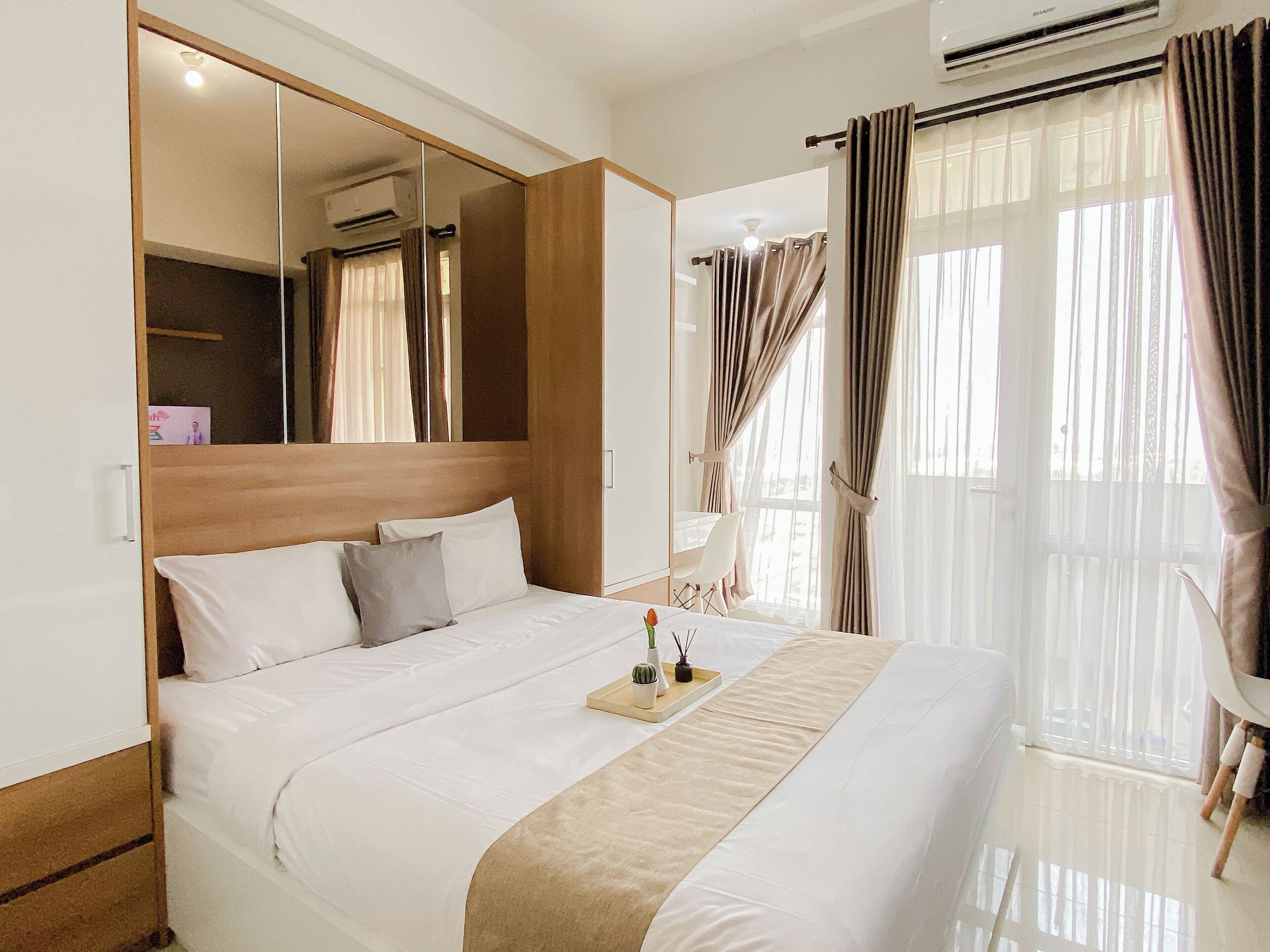 A hotel room with a large bed, a bedside table, a mirror, a chair, and a window with curtains.
