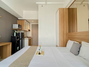 A hotel room with a bed, bedside table, and a kitchenette area with a microwave, refrigerator, and oven. - Warm And Minimalist Studio Apartment Vasanta Innopark (Cikarang)