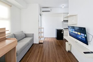 A modern living room with a grey sofa, wooden flooring, a mounted TV, and a small kitchen area with a stove. - Peaceful And Restful Studio Room At Apartment Urbantown Serpong (Tangerang)
