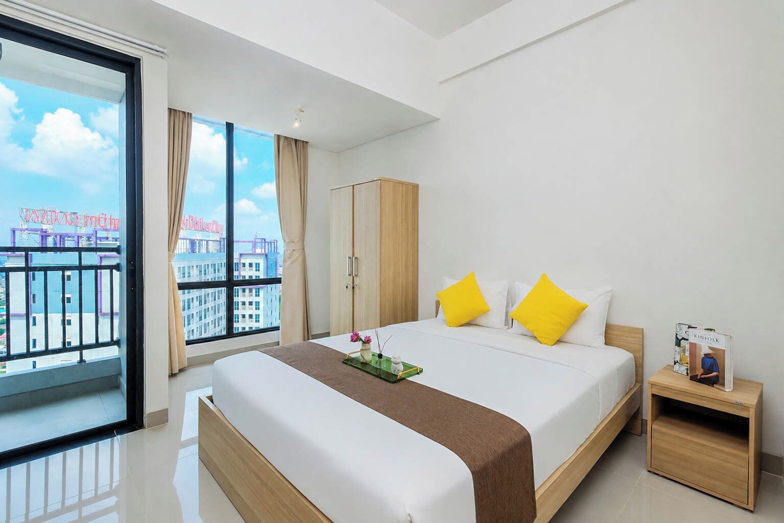 A hotel room with a large window, a bed with white bedding and yellow pillows, a wooden wardrobe, and a nightstand with a book.