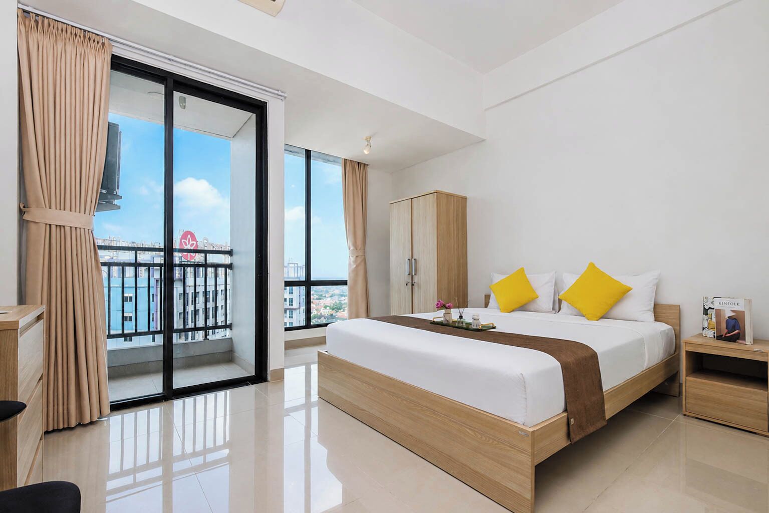 A modern hotel room with a large bed, a wardrobe, a bedside table, and a view of the city through floor-to-ceiling windows.