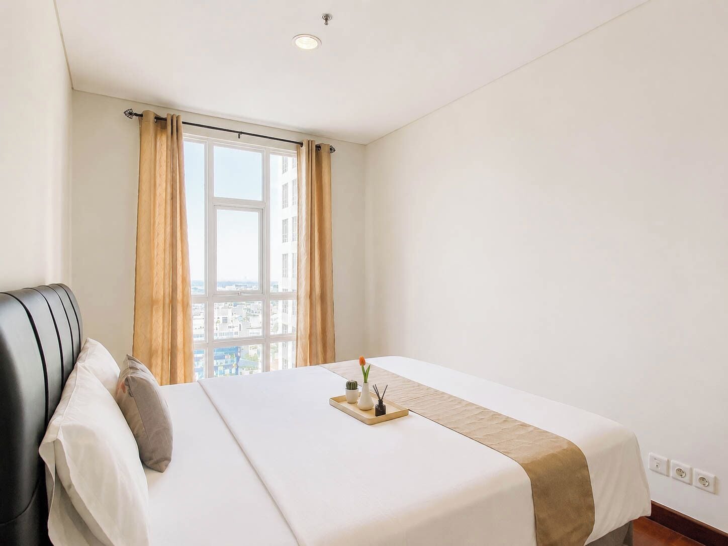 Habitación de hotel con cama, ventana con cortinas, vista a la ciudad y mesita de noche con bandeja.