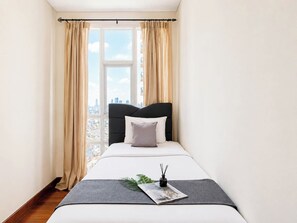 A bedroom with a bed, curtains, a window with a city view, and a small table with a magazine and a plant. - Serene And Spacious 3Br At Apartment Lucky Tower Residence (Jakarta)