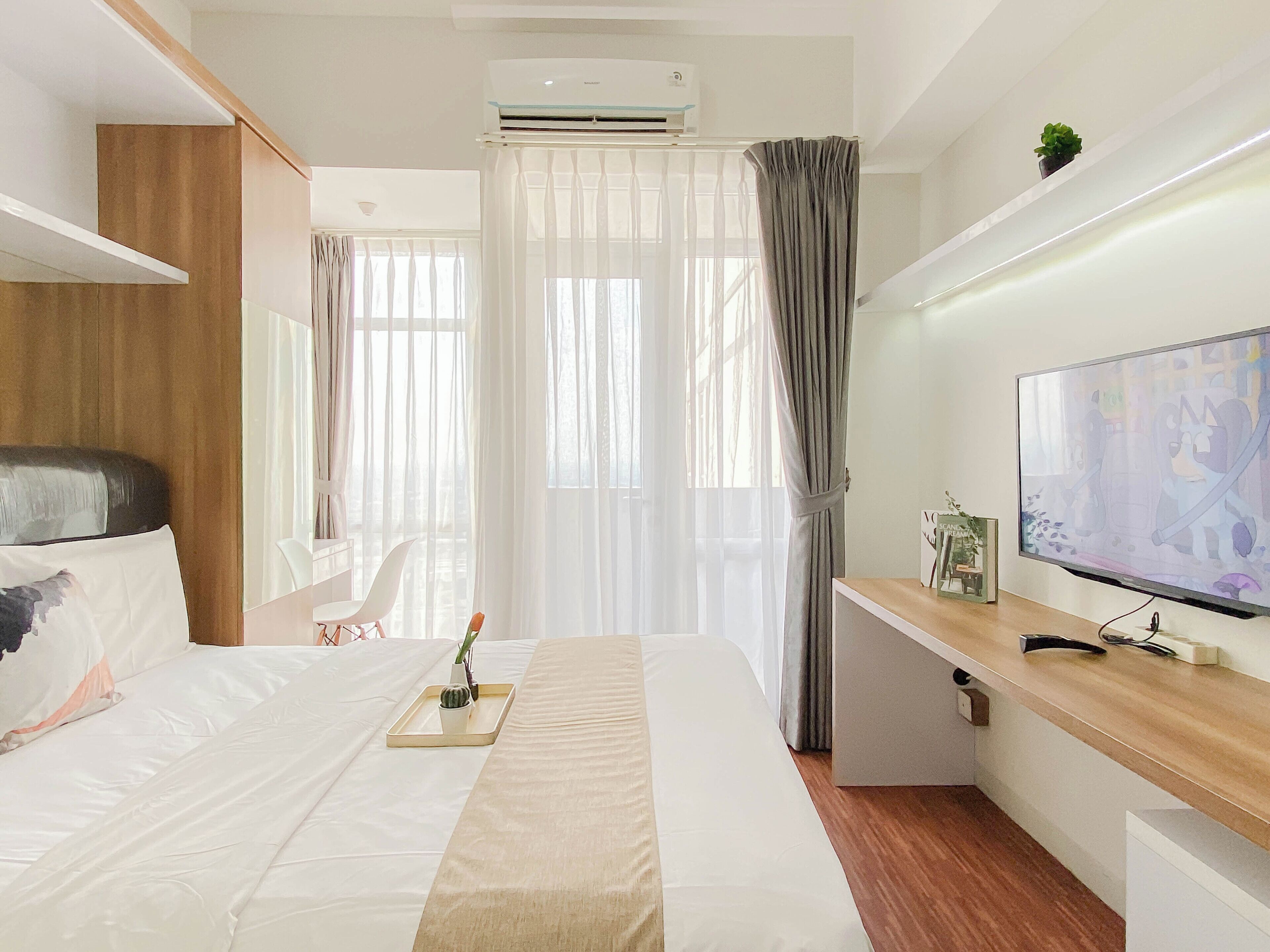 A hotel room with a bed, a desk with a flat-screen TV, an air conditioning unit, and a window with curtains.