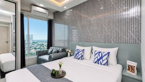 A modern hotel room with a large bed, a seating area with a sofa, a view of the city, and an air conditioning unit. - Calm Living In Gray Studio Room At Apartment Grand Kamala Lagoon (Bekasi)