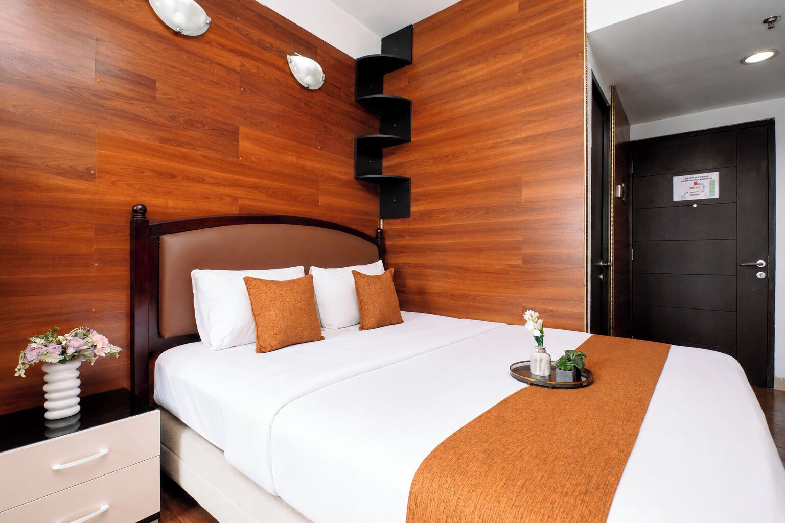 Een hotelkamer met een houten hoofdeinde, een bed met wit beddengoed en twee bruine kussens, een nachtkastje met een bloevas, en een dienblad met een plant en een kaars.