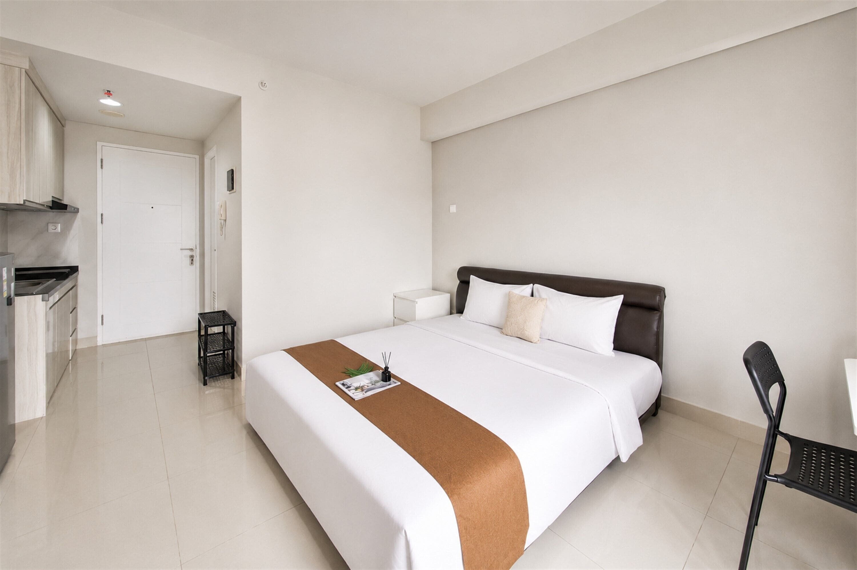 A hotel room with a bed, a bedside table, a chair, and a kitchenette area.