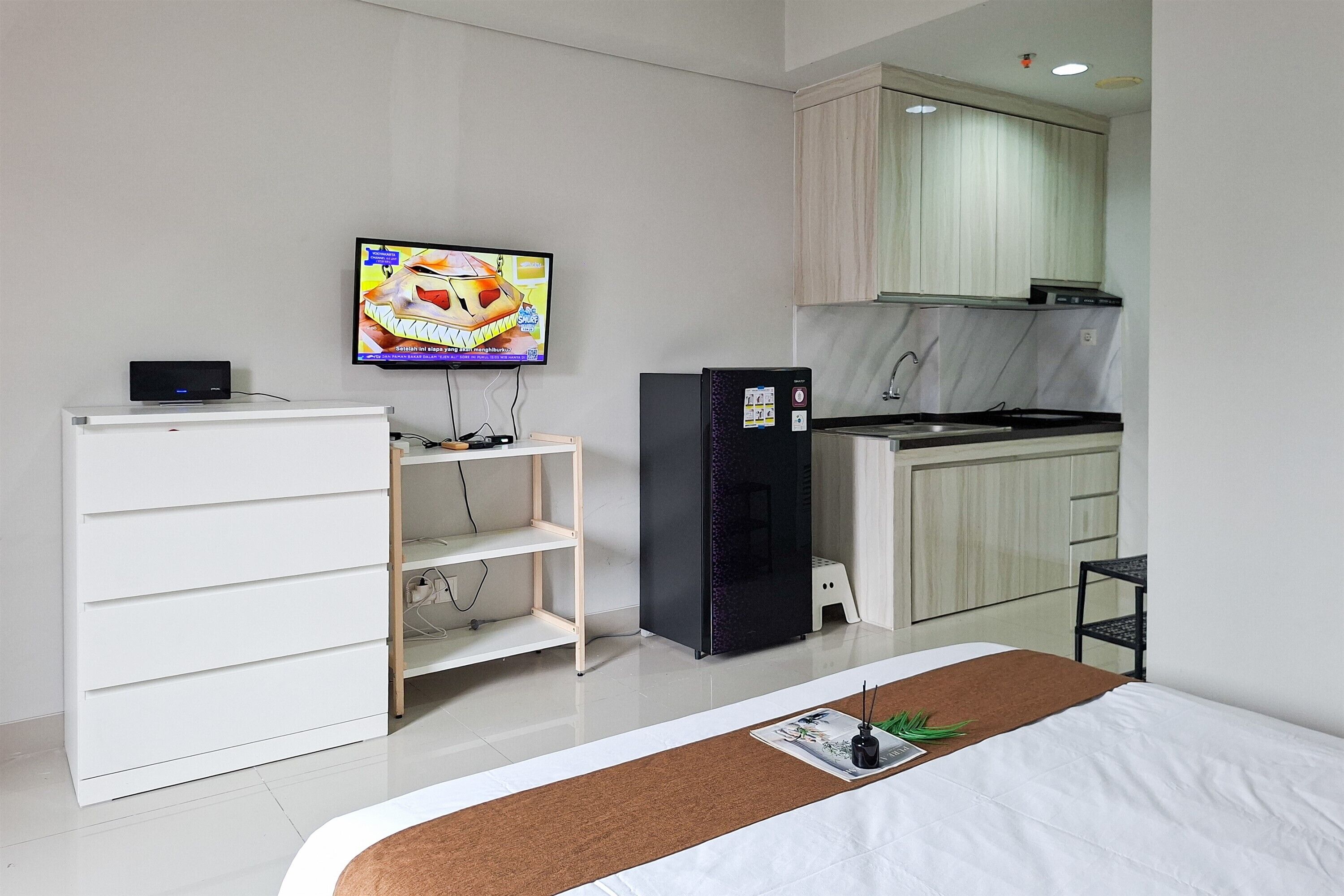 A compact hotel room with a kitchenette, a television, a white cabinet, and a bed with a brown headboard.