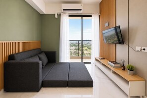 A modern living room with a sofa, a flat-screen TV, a wooden panel wall, and a view of the city. - Chic Fully Furnished 2Br At Apartment Sky House Alam Sutera (Tangerang)