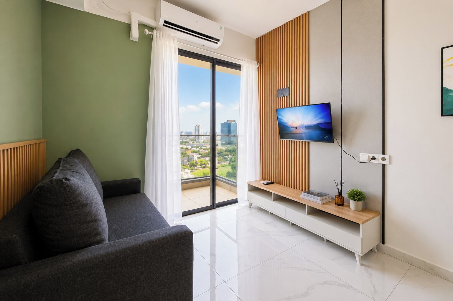 A modern living room with a grey sofa, a flat-screen TV mounted on a wooden panel, and a balcony with a city view.