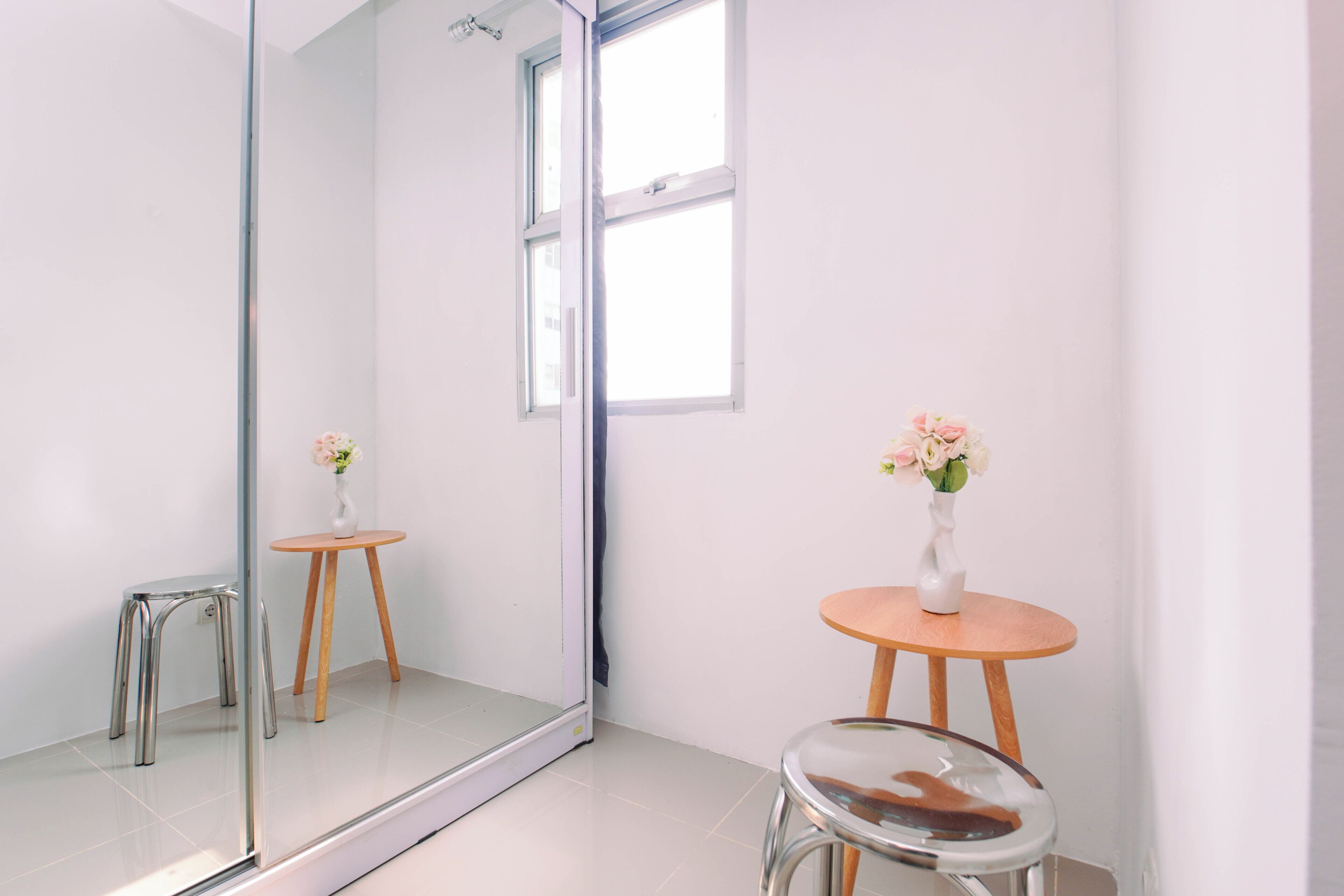Una habitación minimalista con un espejo grande, un taburete pequeño y una mesa con un jarrón de flores.