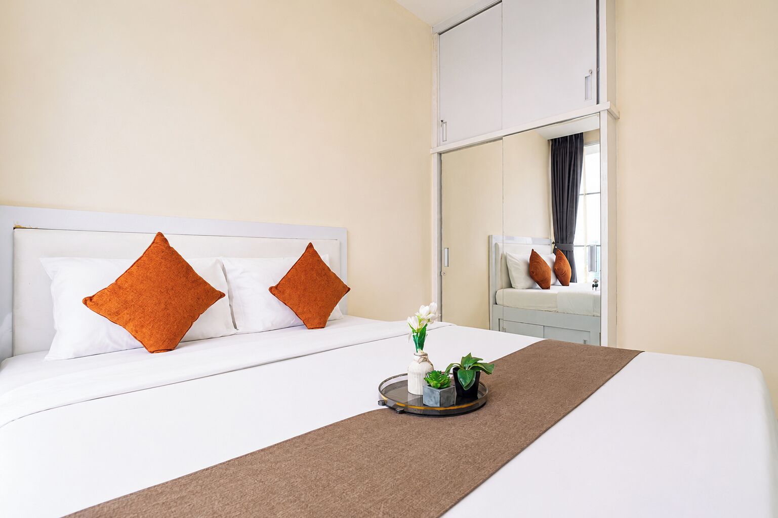 A hotel room with a bed, pillows, a bedside table with a vase and plants, and a mirror.