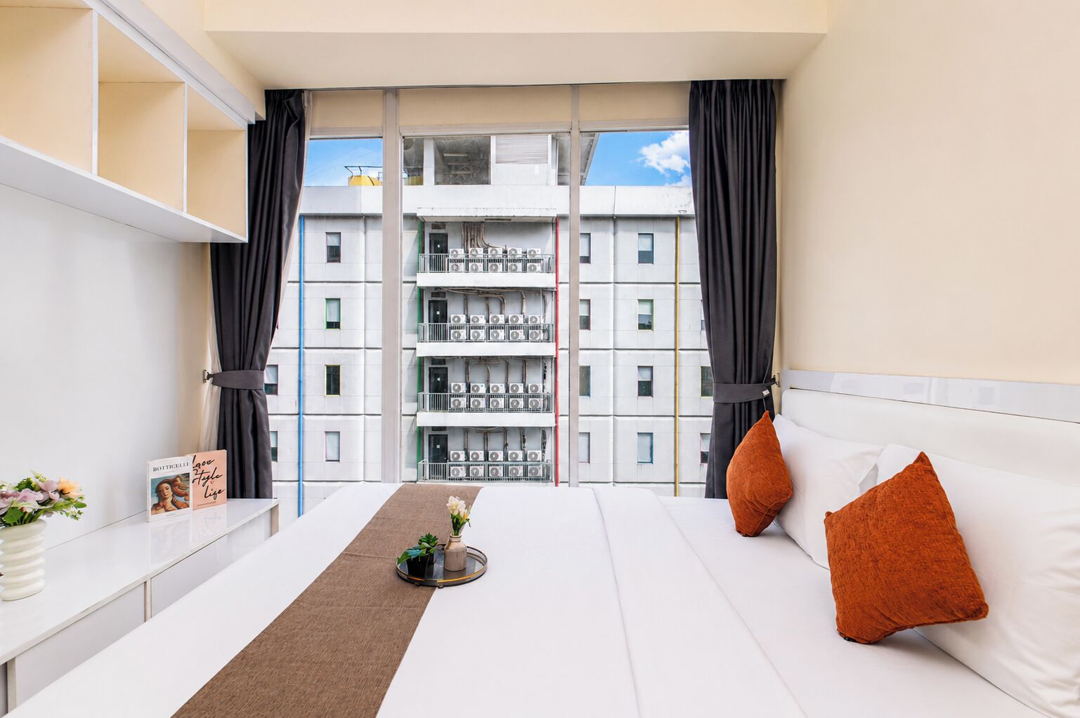 A hotel room with a bed, a bedside table, a window with curtains, and a view of a building with multiple floors and balconies.