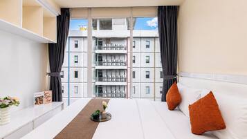 A hotel room with a bed, a bedside table, a window with curtains, and a view of a building with multiple floors and balconies.