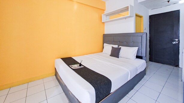 Quarto de hotel com cama, mesa de cabeceira, prateleira na parede e porta.