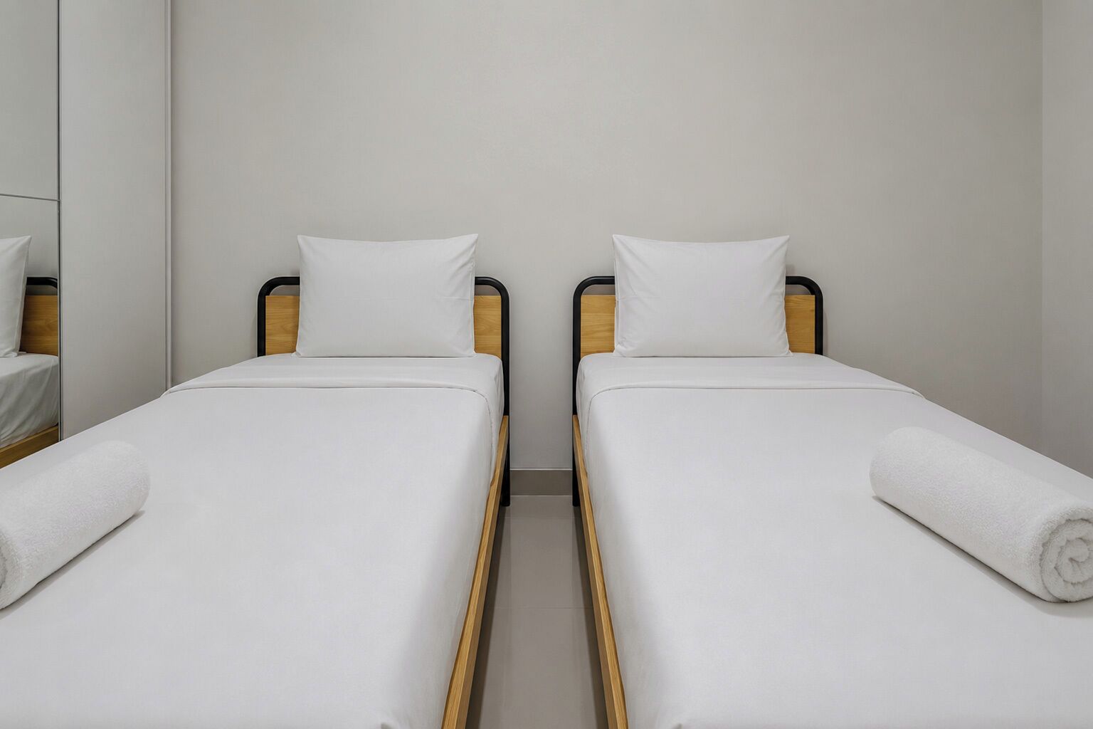 Deux lits simples avec des cadres en bois et du linge de lit blanc, dans une pièce aux murs unis et dotée d’un miroir.
