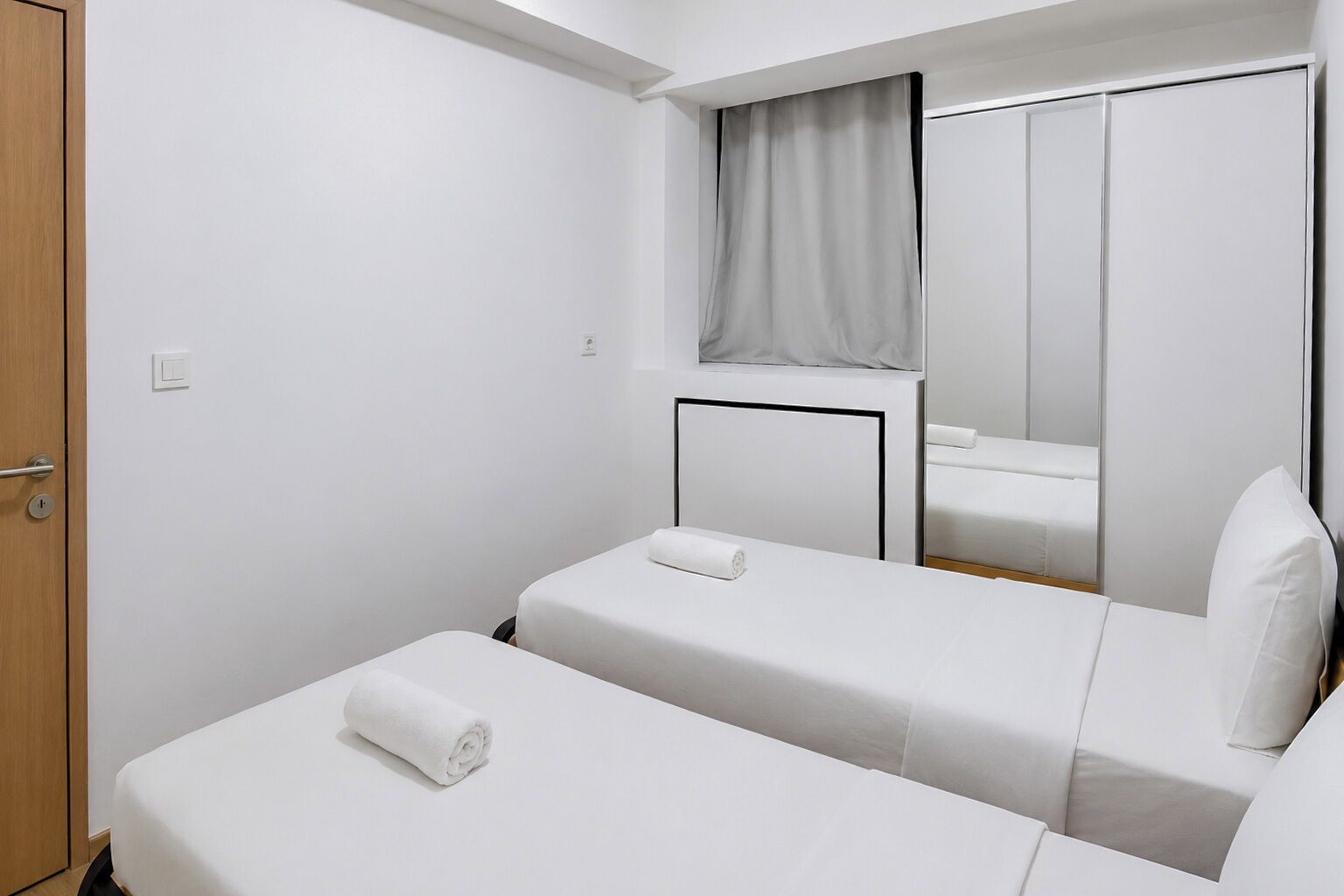 Habitació d'hotel amb dos llits individuals, una porta de fusta, un mirall i una finestra amb cortina.