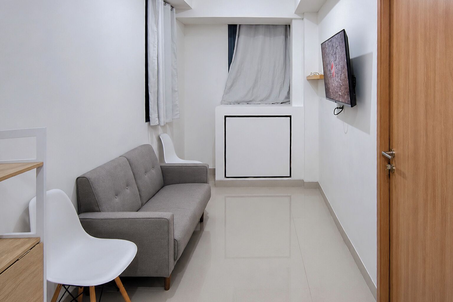 Ein kleiner, moderner Raum mit einem grauen Sofa, einem weißen Sessel, einem an der Wand befestigten Fernseher und einer Holztür.