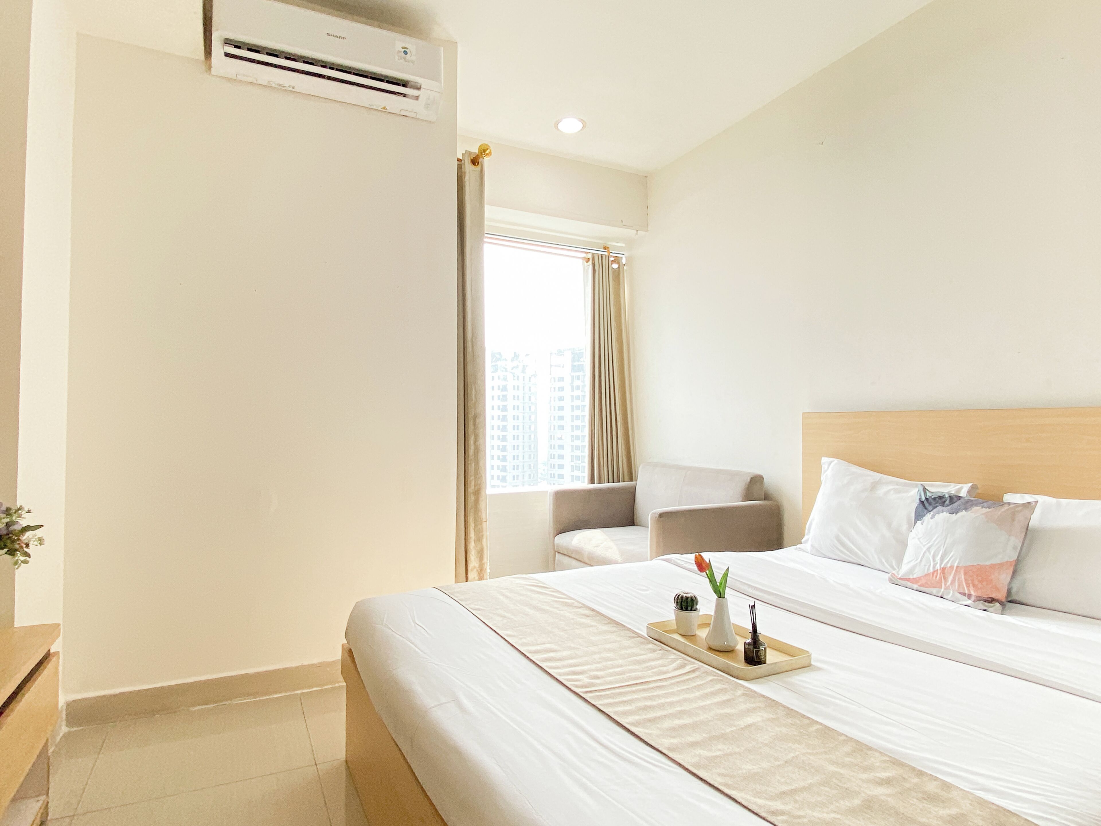 A hotel room with a bed, a chair, a window with curtains, an air conditioning unit, and a tray with a vase and a plant.