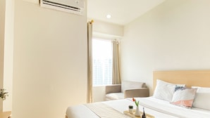 A hotel room with a bed, a chair, a window with curtains, an air conditioning unit, and a tray with a vase and a plant. - Minimalist Functional Studio At Apartment Grand Kamala Lagoon (Bekasi)