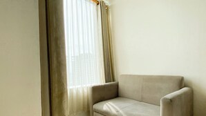 Una habitación de esquina con un sofá color arena, una ventana con cortinas translúcidas y una pared blanca lisa.